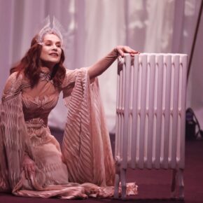 Isabelle Huppert kneeling in a pleated peach gown beside a white radiator in Castellucci’s Bérénice at the 2025 Holland Festival.