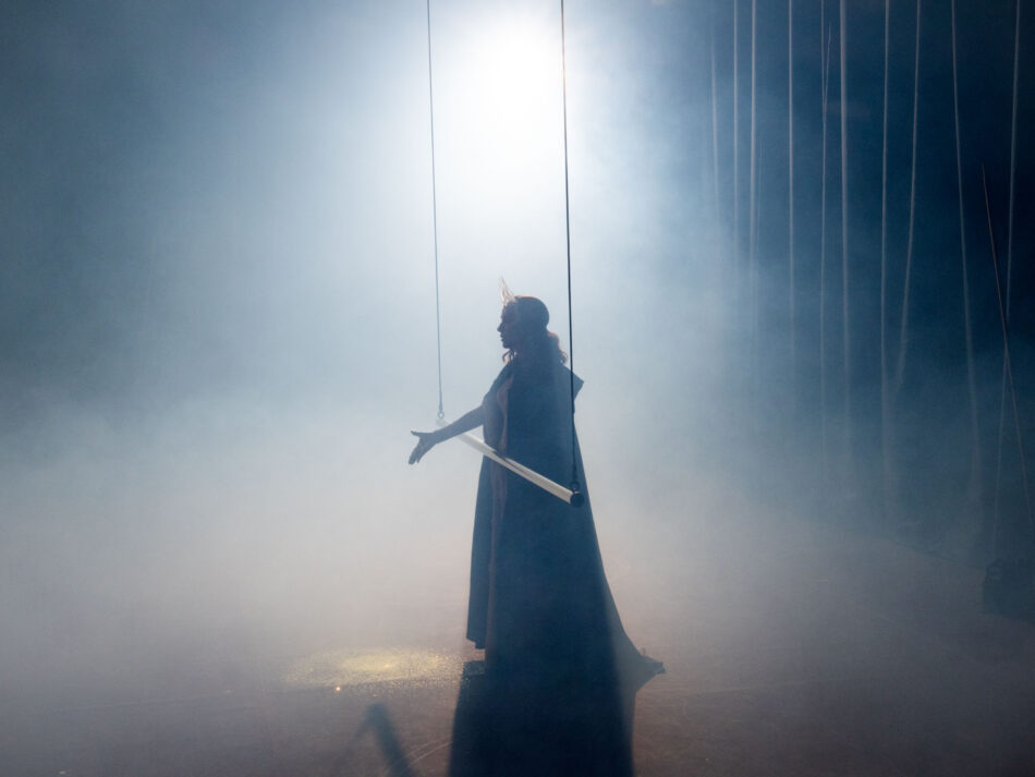 Isabelle Huppert in a silhouetted moment from Romeo Castellucci’s Bérénice at the 2025 Holland Festival, surrounded by mist, suspended rods, and stark theatrical light. © Alex Majoli Silhouette of Isabelle Huppert as Bérénice in Romeo Castellucci’s Holland Festival production, framed in fog with suspended rods and backlight.