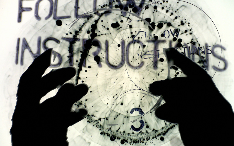 Hands silhouetted against projected text and inked markings, suggesting layered image and gesture during a live performance at The Dark Festival.